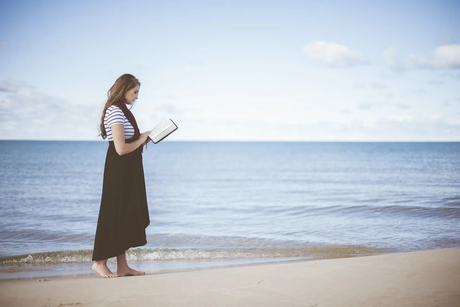 Woman reading a book while walking barefoot along a quiet ocean shoreline